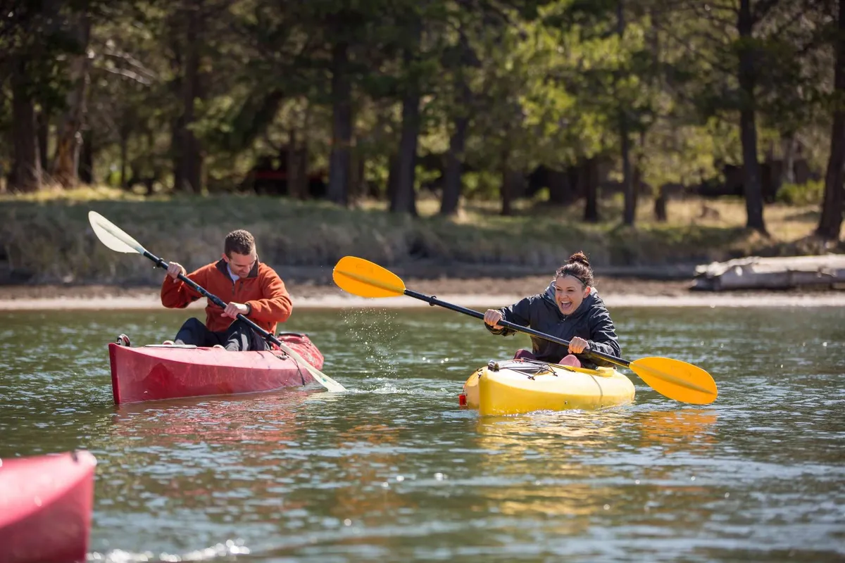 Two people paddle bright, colorful kayaks on a sparkling river, with lush greenery along the banks and their reflections shimmering on the water. The image captures the sense of excitement and companionship that comes from exploring a waterway together.
