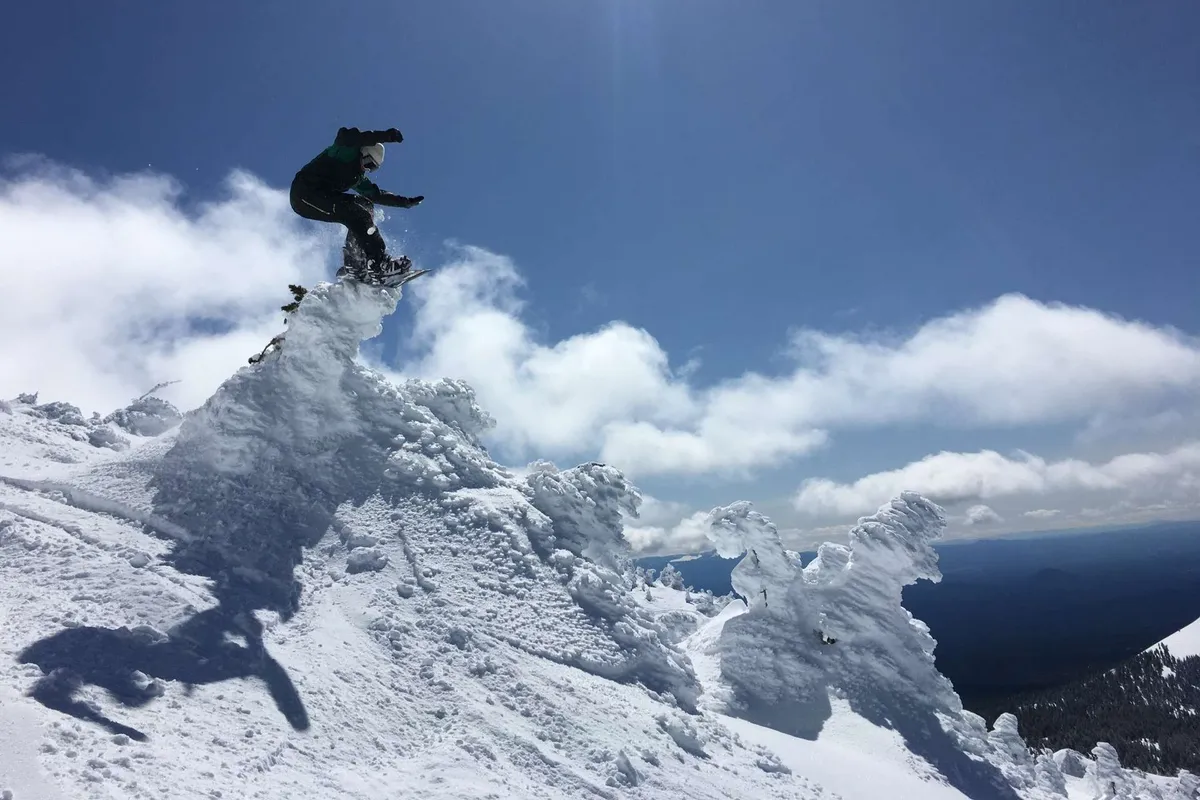 A dynamic action shot of a snowboarder mid-air against a backdrop of a snow-covered peak and expansive clouds. The scene conveys excitement, adventure, and the thrill of exploring Bend's nearby mountains.