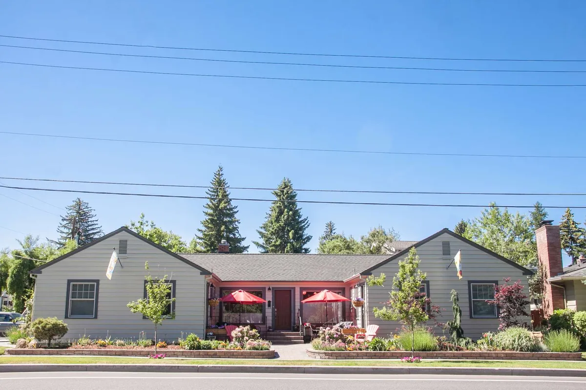 A cheerful exterior shot featuring colorful flower beds, bright red umbrellas, and a welcoming front porch. The scene suggests a relaxed outdoor environment ideal for sunset dining or enjoying the neighborhood atmosphere.