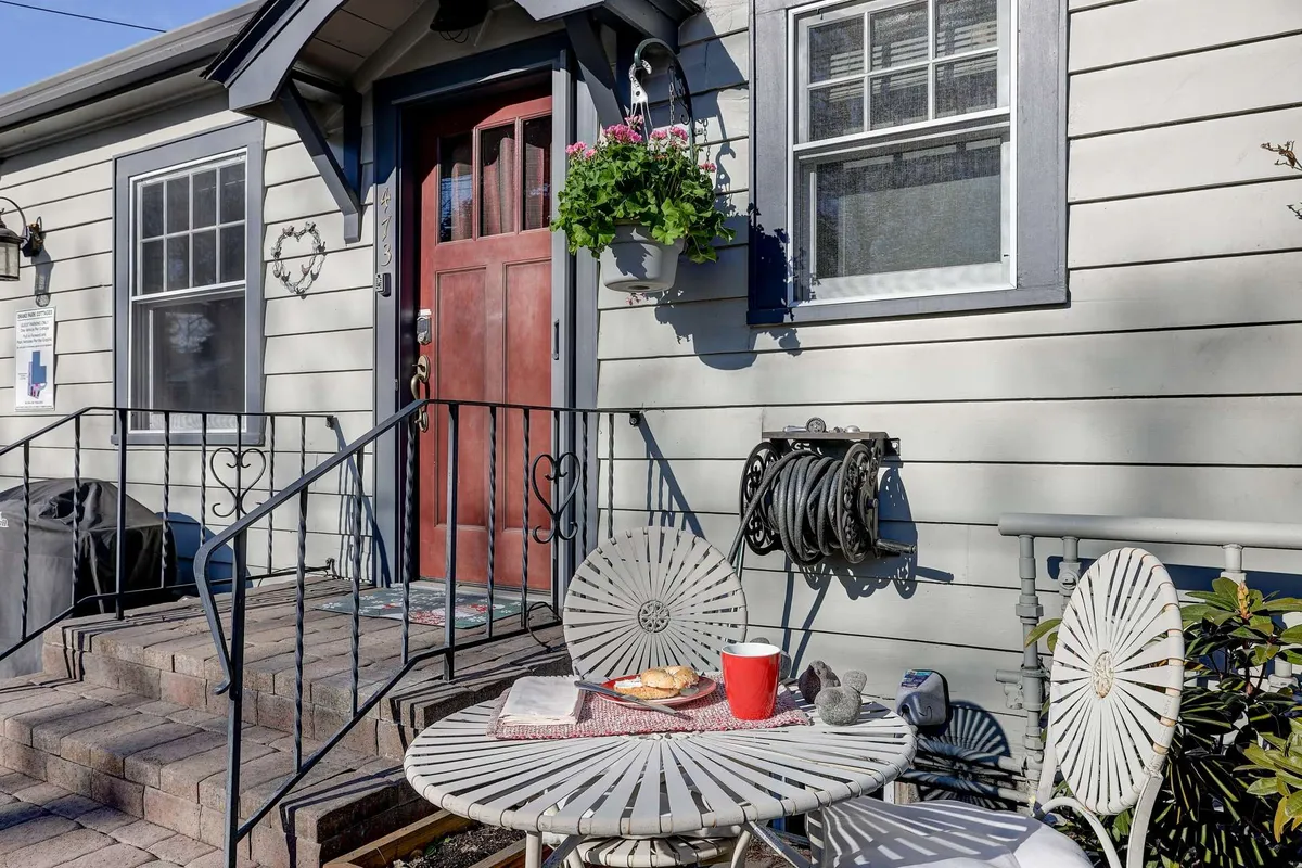 A charming exterior view with outdoor seating by the front door, complemented by a potted plant. The space radiates coziness and is staged for morning coffee or intimate gatherings, with subtle hints of surrounding foliage.