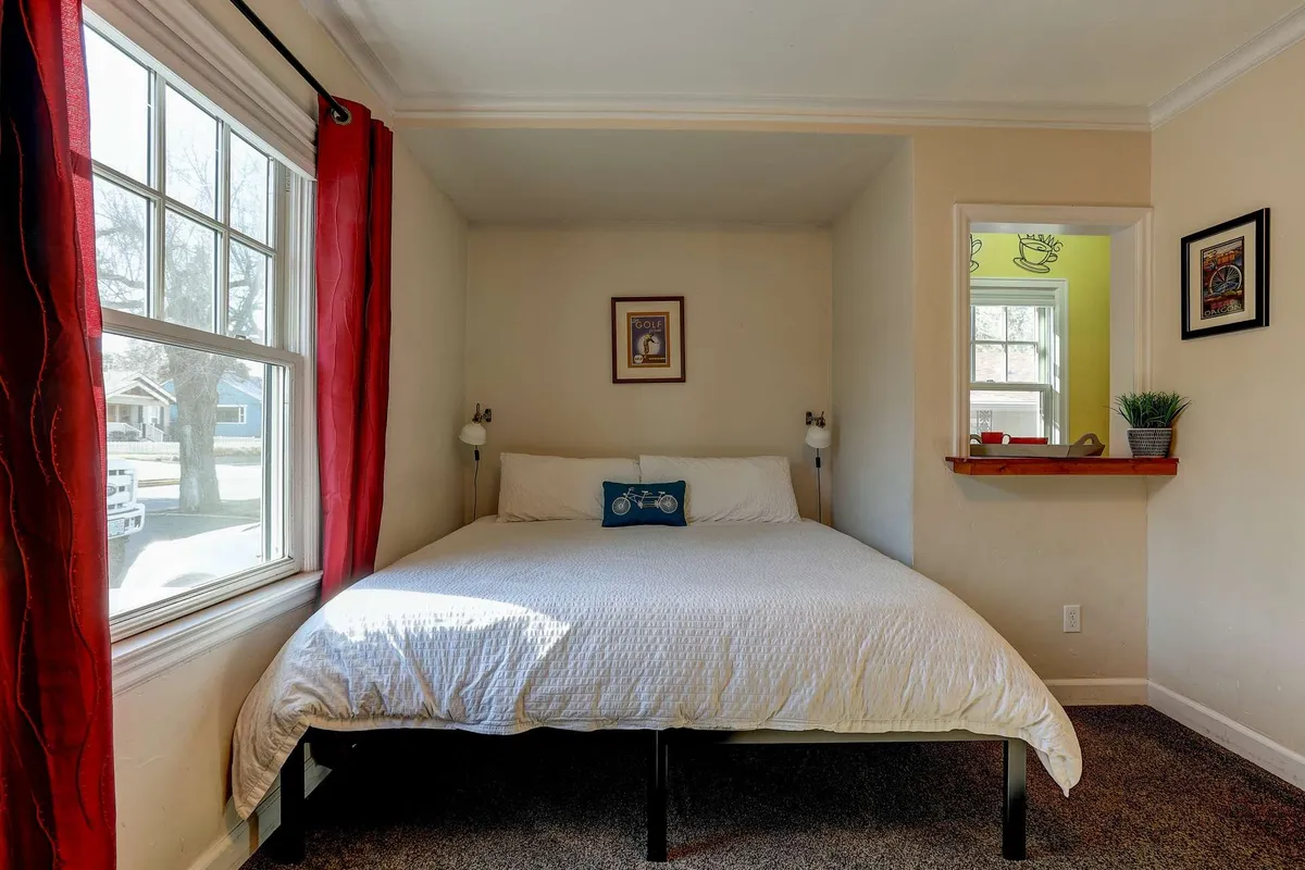 A serene sleeping area with a comfortable bed, a window letting in natural light, a decorative frame, and a shelf with a plant. The ambiance is restful and inviting, with touches that suggest both relaxation and thoughtful design.