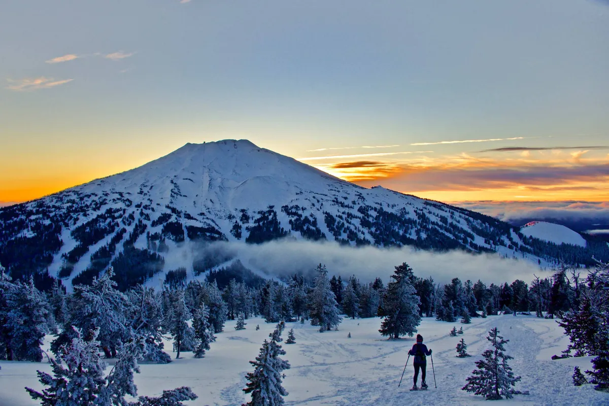 A breathtaking exterior view of a snow-covered mountain at sunset, framed by snowy trees, with a skier in the foreground. The tranquil landscape evokes the beauty and grandeur of Central Oregon's natural surroundings.
