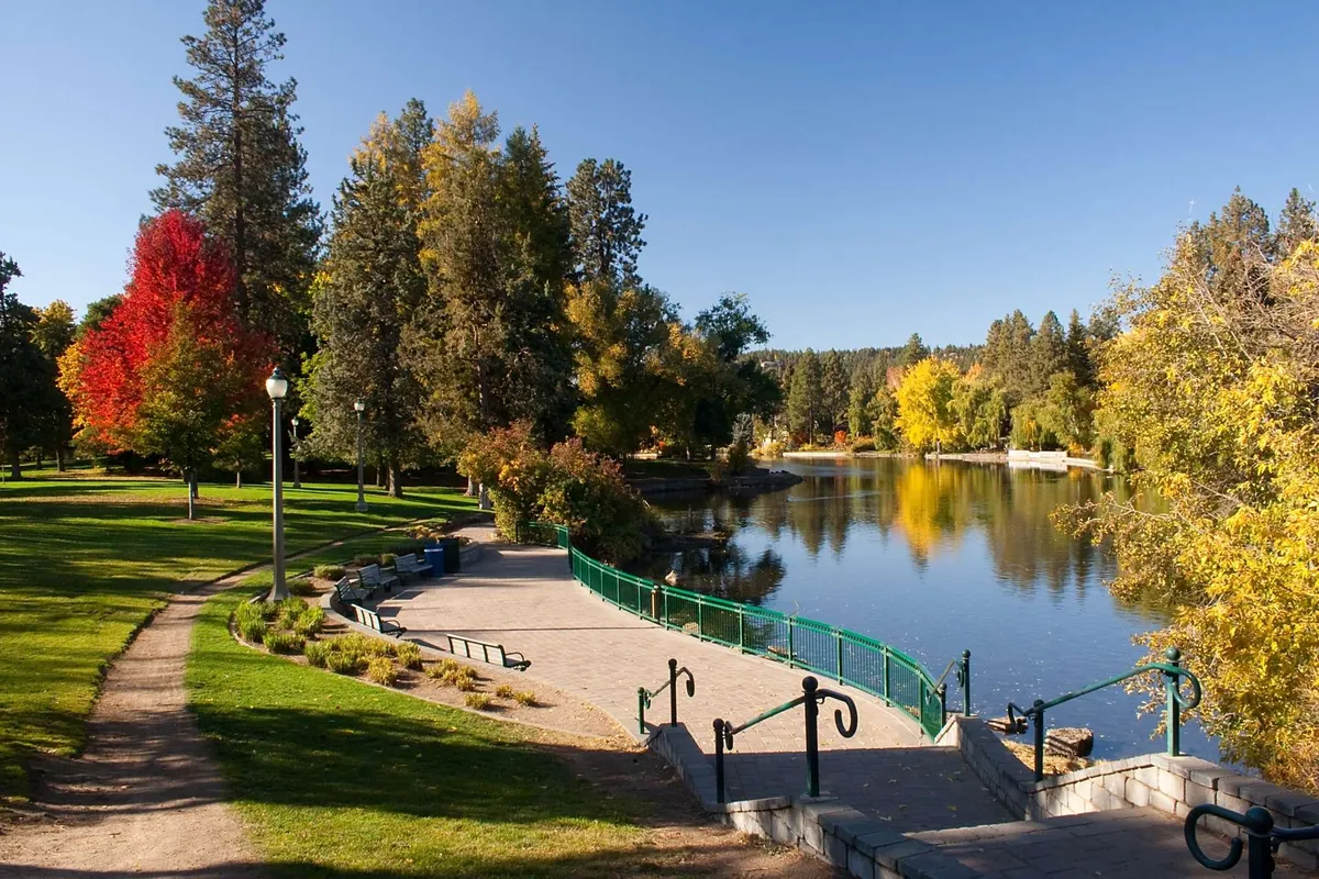 A tranquil view of a tree-lined lake with a pathway, benches, and a bridge, inviting visitors to walk, relax, and soak in the natural beauty. The environment hints at a vibrant community space, suitable for picnics, casual strolls, and enjoying the park's atmosphere, all just steps from the residence.