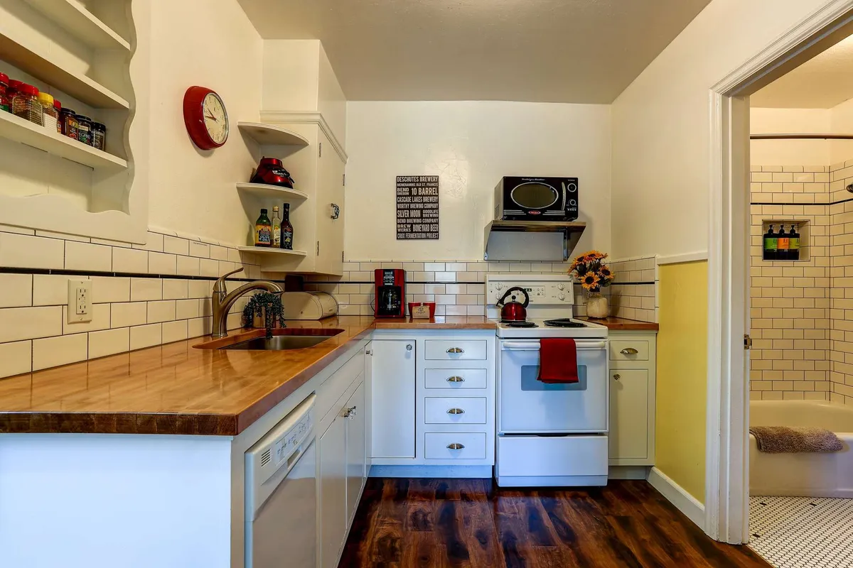 A bright, welcoming kitchen scene featuring a kitchen countertop with a visible stove, shelves lined with spices, a decorative clock, and a flower vase. The layout feels functional and cozy, emphasizing the inviting mood of meal preparation and sharing.