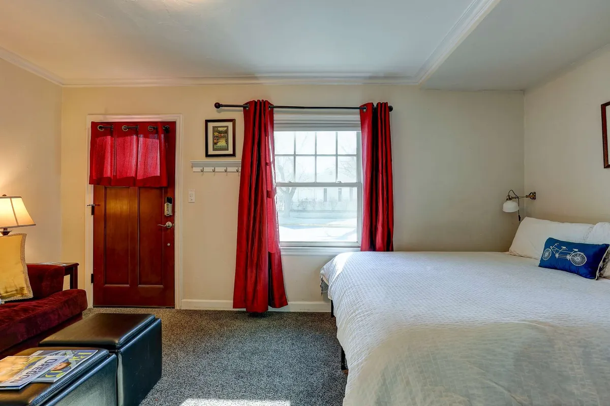 A welcoming studio interior with a cozy bed, red curtains, wooden door, and a comfy chair by the window. Sunlight fills the space, inviting guests to unwind after a day of adventure outside.