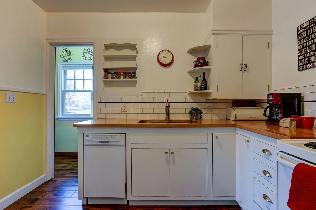 A bright, inviting kitchen featuring a wooden countertop, open shelving with dishware, a retro wall clock, and modern appliances. Sunlight fills the space, and the countertop is set for a relaxed morning coffee or a creative work session.