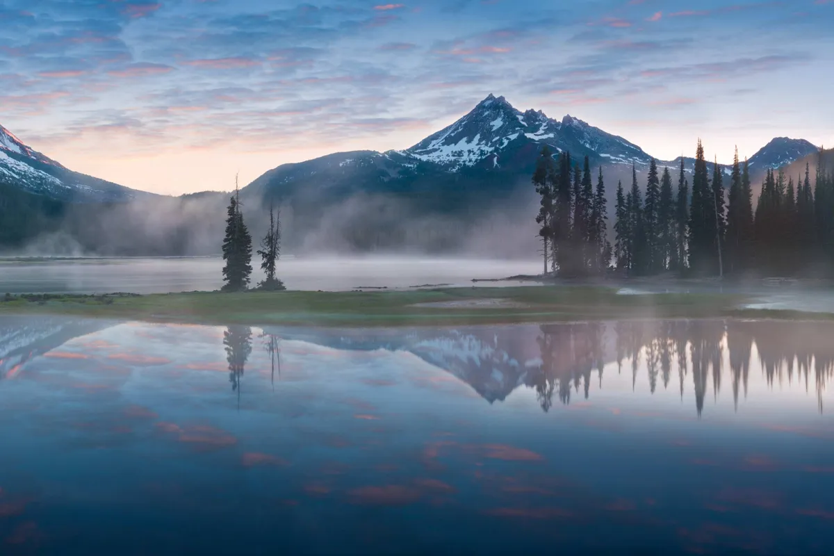 A peaceful lakeside view with mountains, reflective water, and tree-lined shores. The natural beauty is enhanced by the soft light, ideal for hiking, relaxing, or sharing quiet moments at the water’s edge.