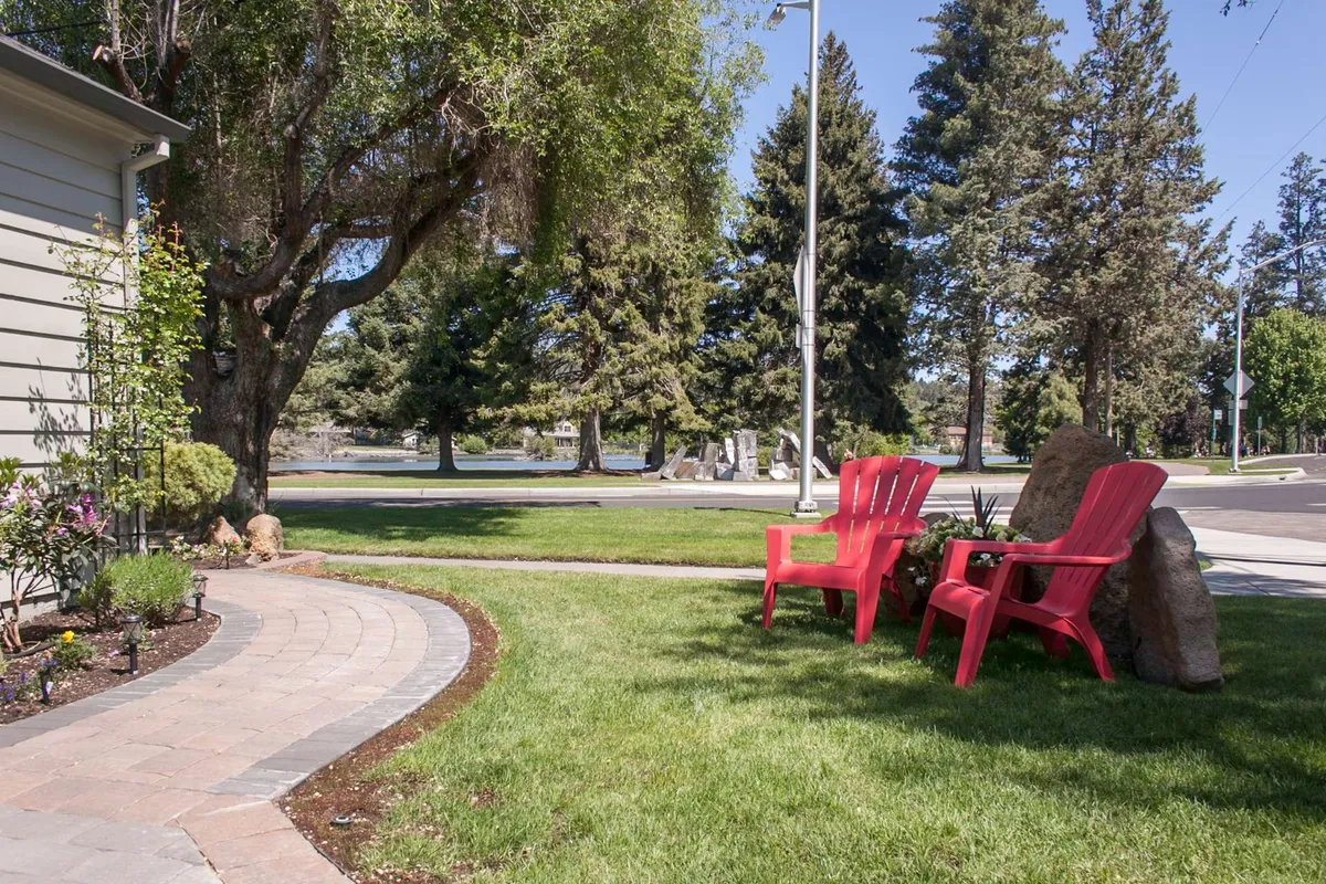 This image shows a cozy outdoor patio area with two red chairs set on a pathway overlooking green grass and mature trees. The setting exudes tranquility and invites relaxing moments, whether it's enjoying a morning coffee or winding down at sunset. The chairs face outward, suggesting a view and connection to surrounding greenery, possibly a park.