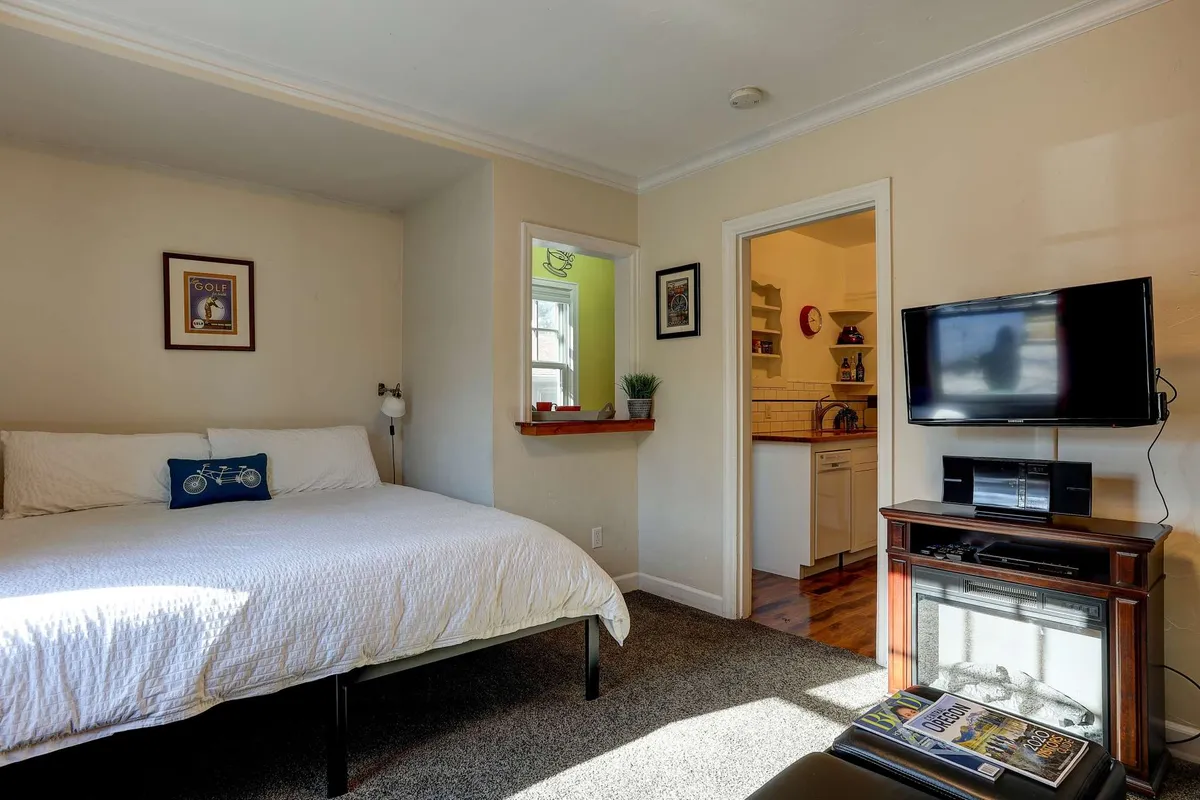 A cozy bedroom with crisp white linens on the bed, modern wall art, a mounted TV, and natural light streaming in. The scene suggests a tranquil spot for reading, remote work, or relaxing after exploring the neighborhood.